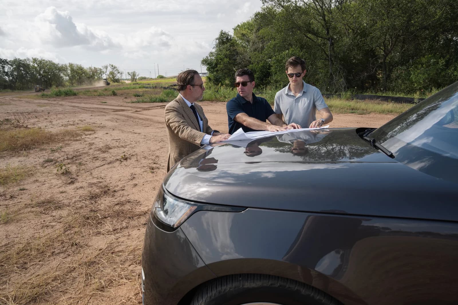 Three people reviewing site plans spread across the hood of a vehicle at the edge of the site