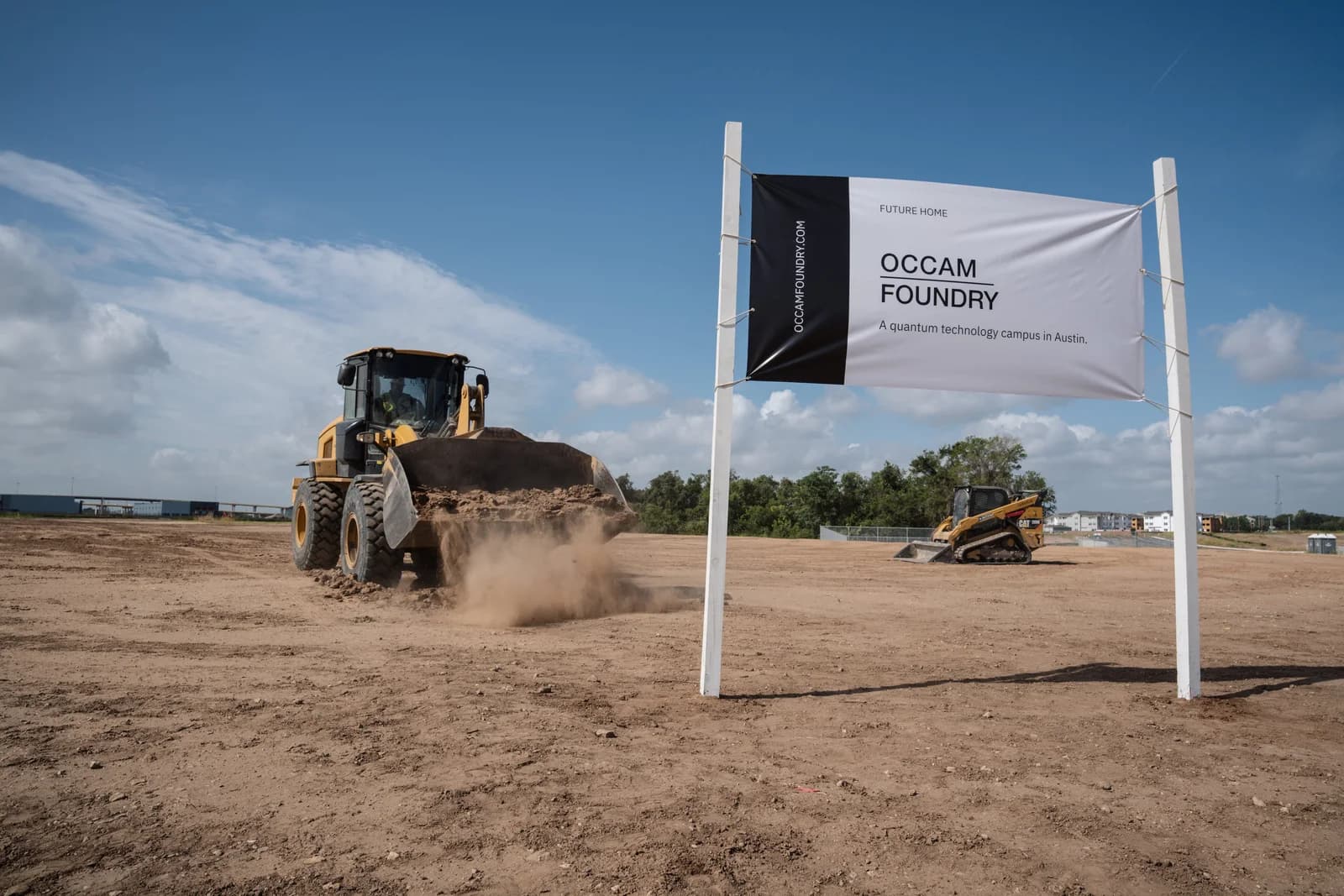 The Occam Foundry site banner standing in the open ground, a wheel loader moving earth behind it